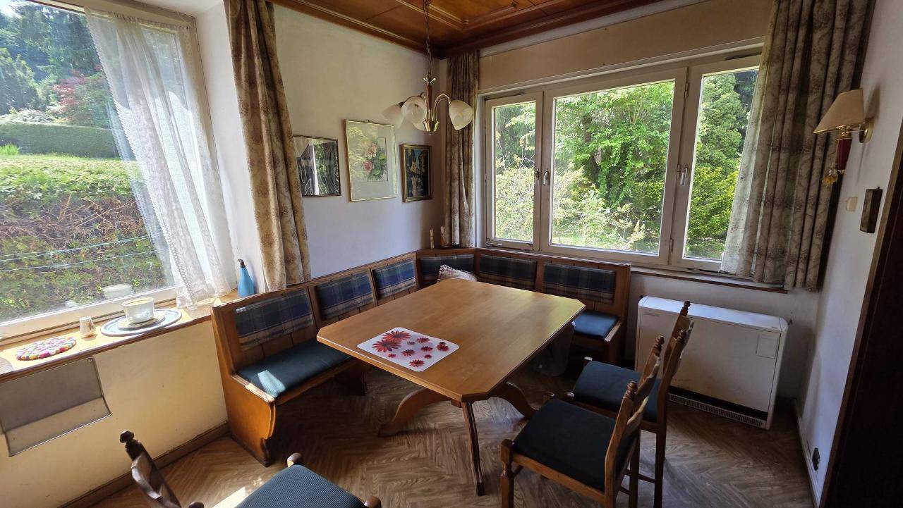 Gemütlicher Essbereich mit Eckbank und Holztisch, umgeben von großen Fenstern mit Blick ins Grüne.