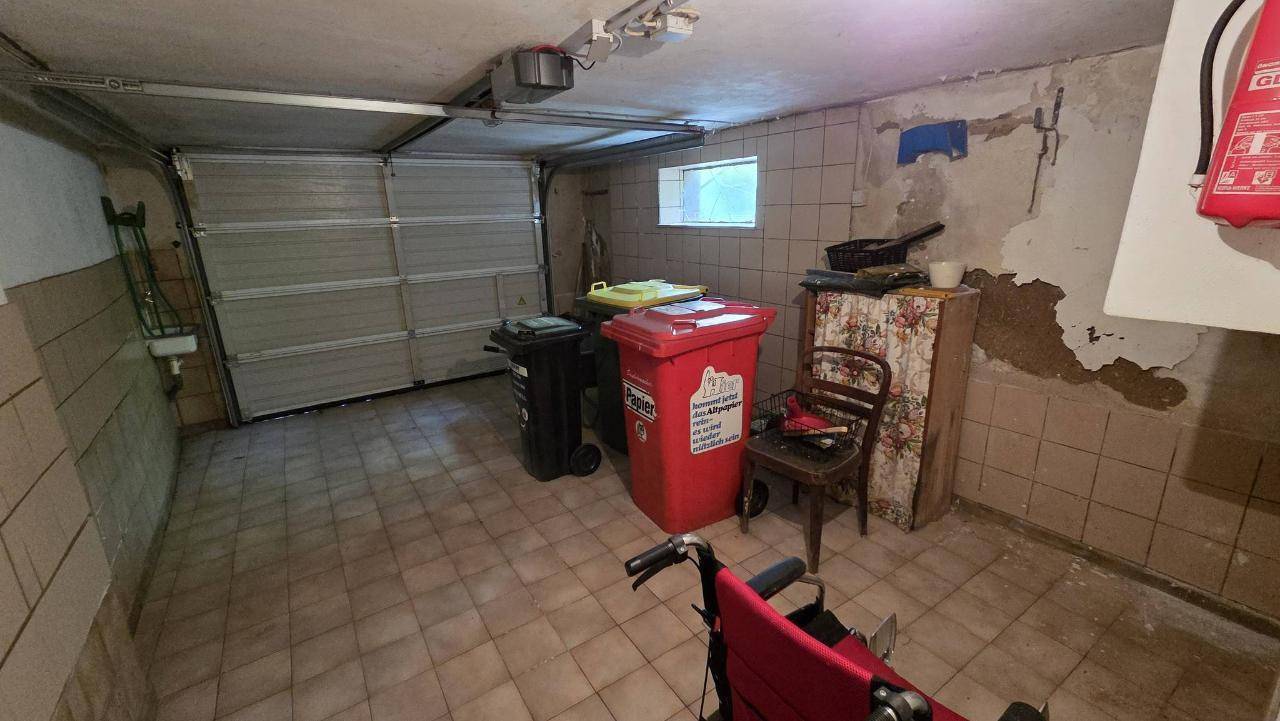 Geräumige Garage mit Fliesenboden, Rolltor und Platz für Fahrzeuge sowie Abfallbehälter.
