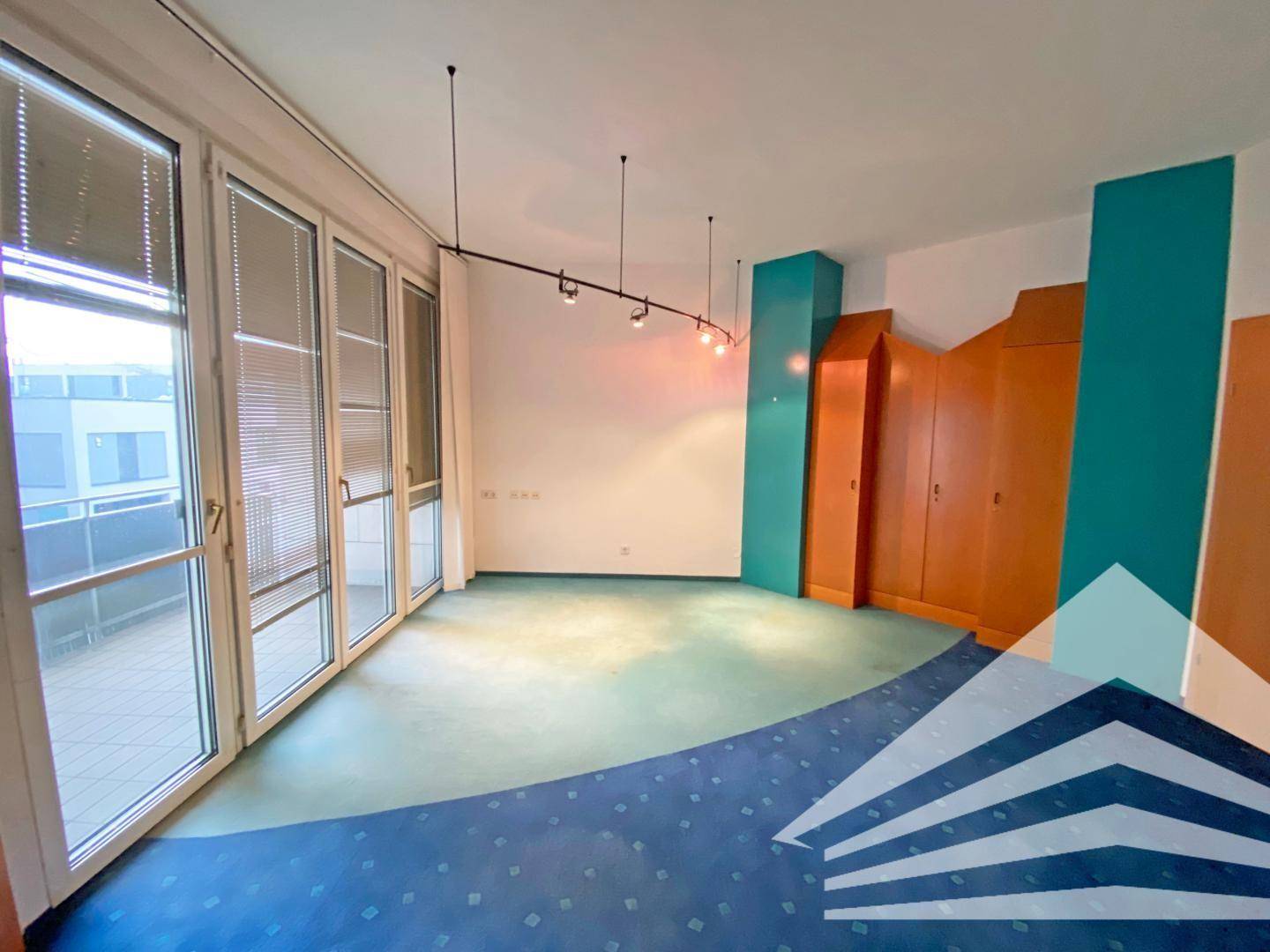 Geräumiges Büro mit großen Fenstern, Zugang zu einem Balkon und einem farbenfrohen Teppichboden.