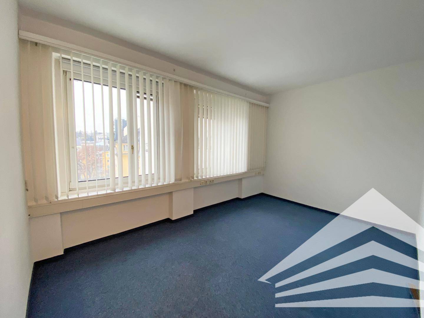 Leeres Büro mit großen Fenstern, vertikalen Jalousien und blauem Teppichboden, ideal für Arbeitsbereiche.