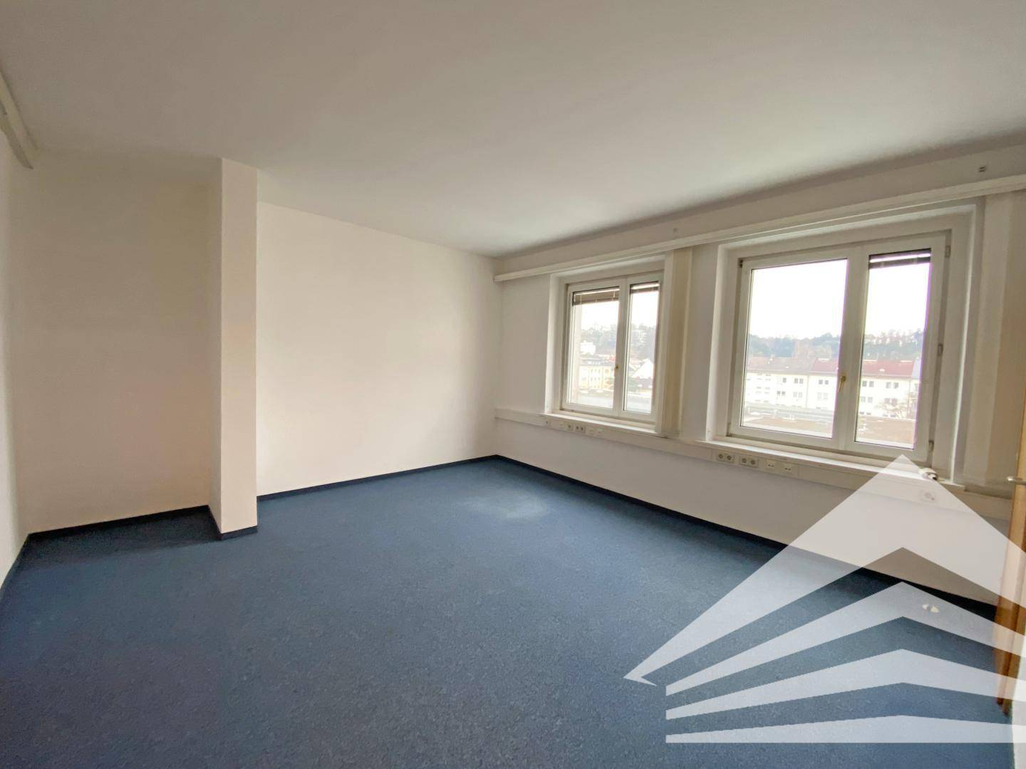 Helles Büro mit zwei Fenstern, weiß gestrichenen Wänden und einem blauen Teppichboden, bereit zur Einrichtung.
