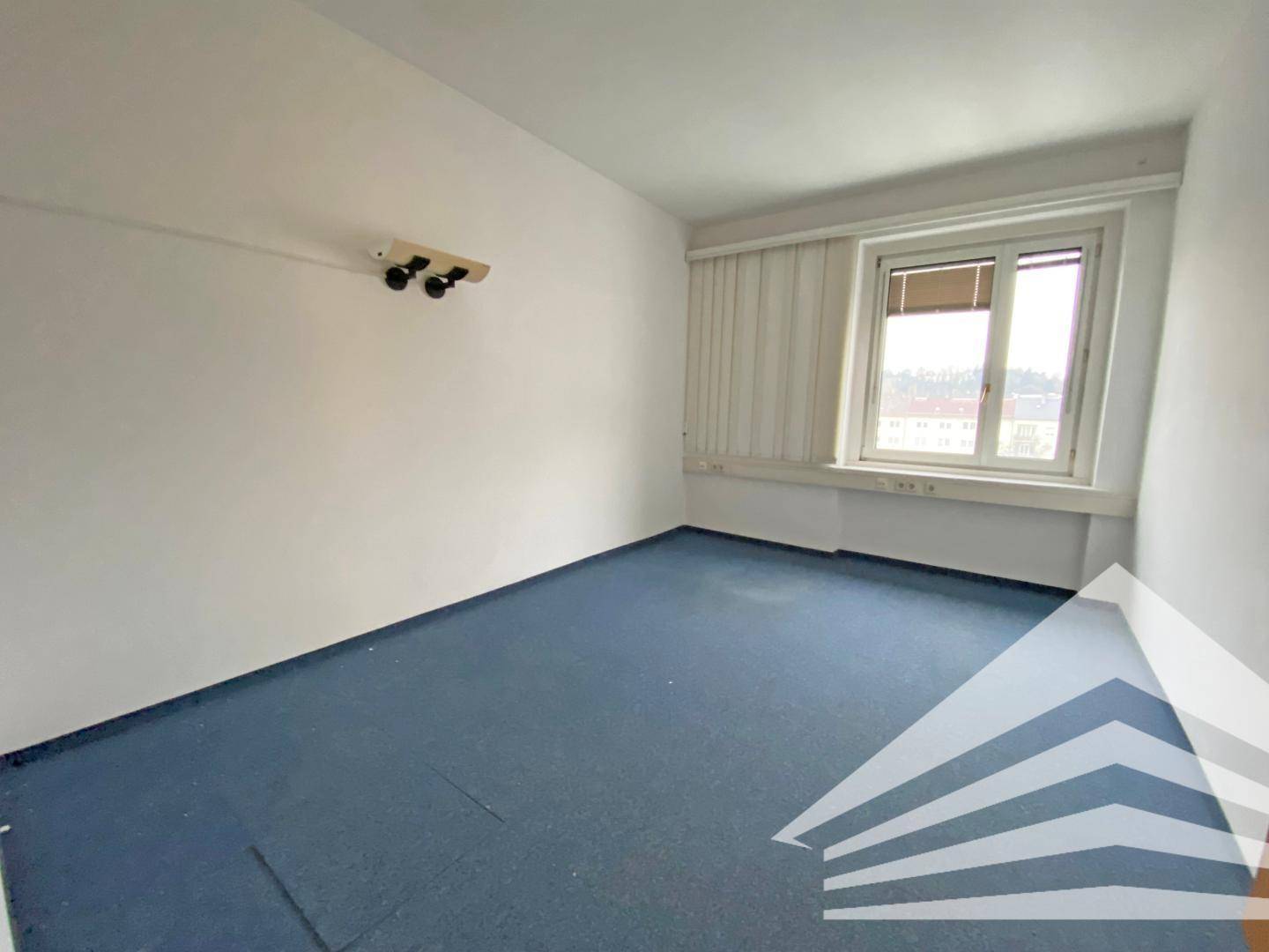 Einfaches Büro mit blauem Teppichboden, einem Fenster mit Jalousien und Wandleuchten für zusätzliche Beleuchtung.