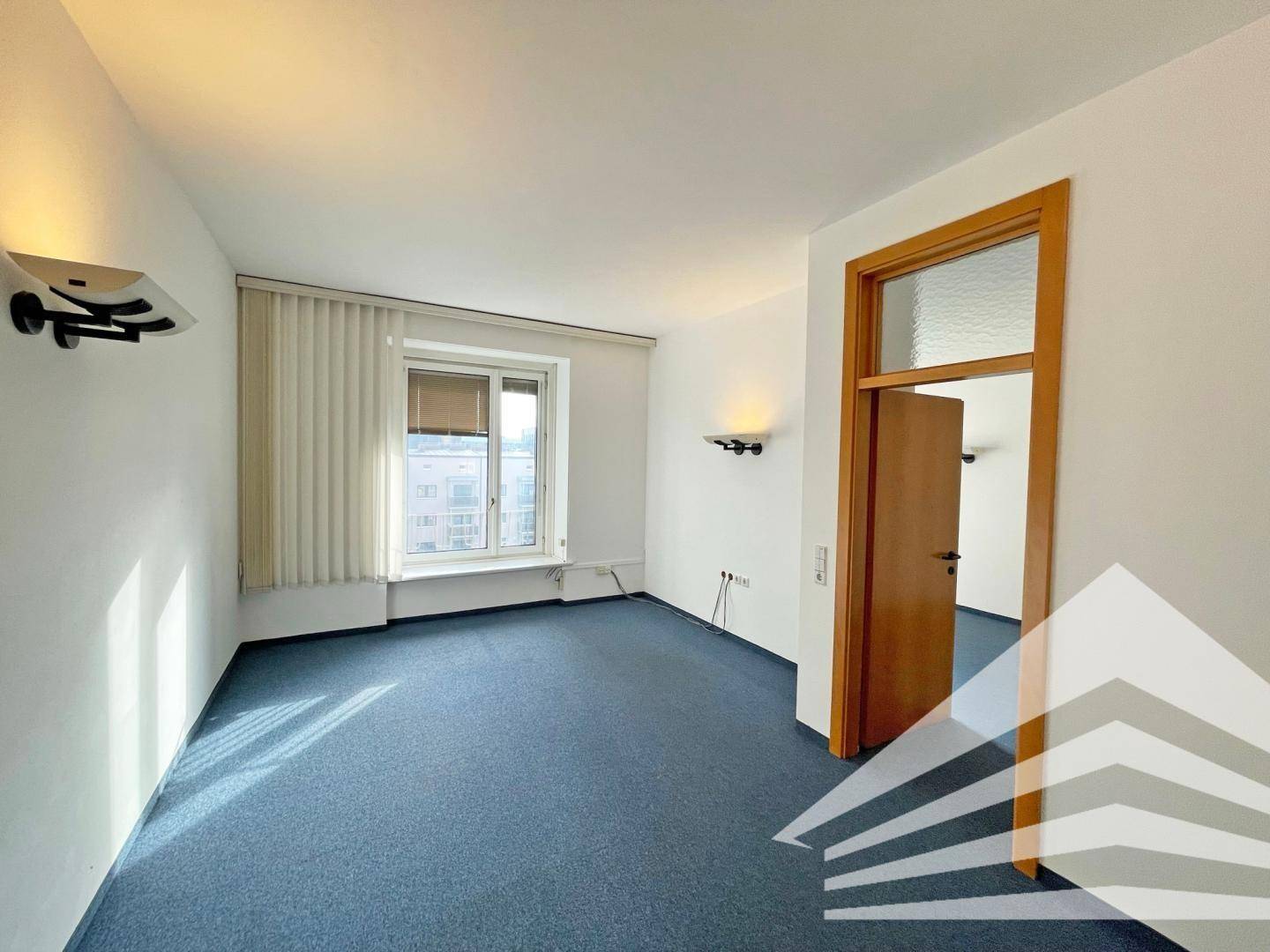 Büro mit blauem Teppichboden, Wandleuchten und einer Holztür, die zu einem weiteren Raum führt.