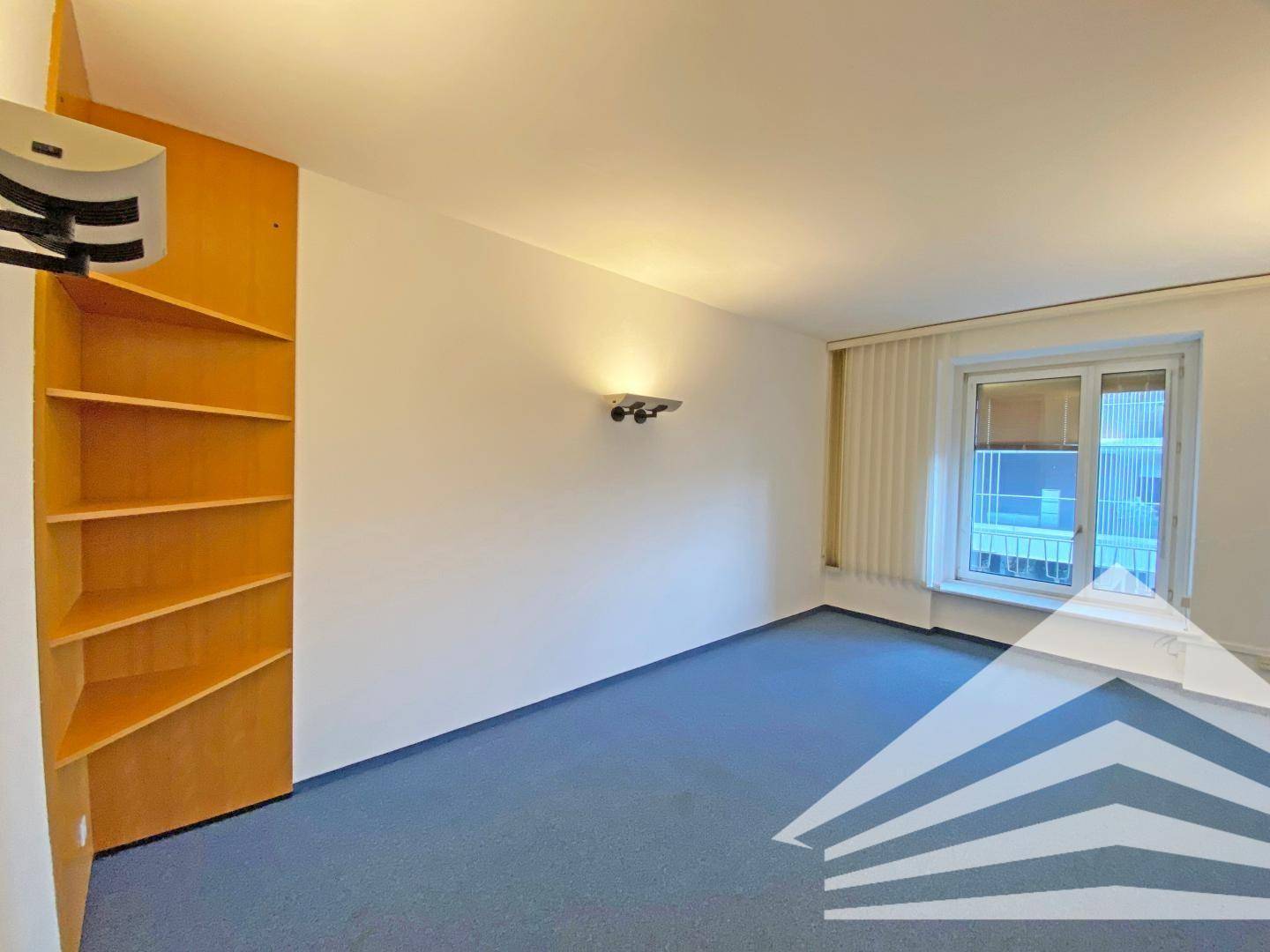 Helles Büro mit blauem Teppichboden, Einbauregal und einem Fenster mit Blick nach draußen.
