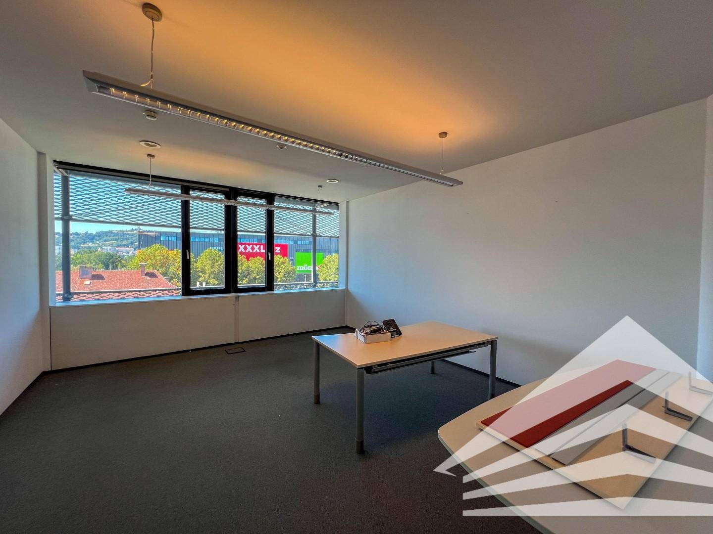 Funktionales Büro mit Teppichboden, einem einfachen Tisch und großen Fenstern mit Jalousien.