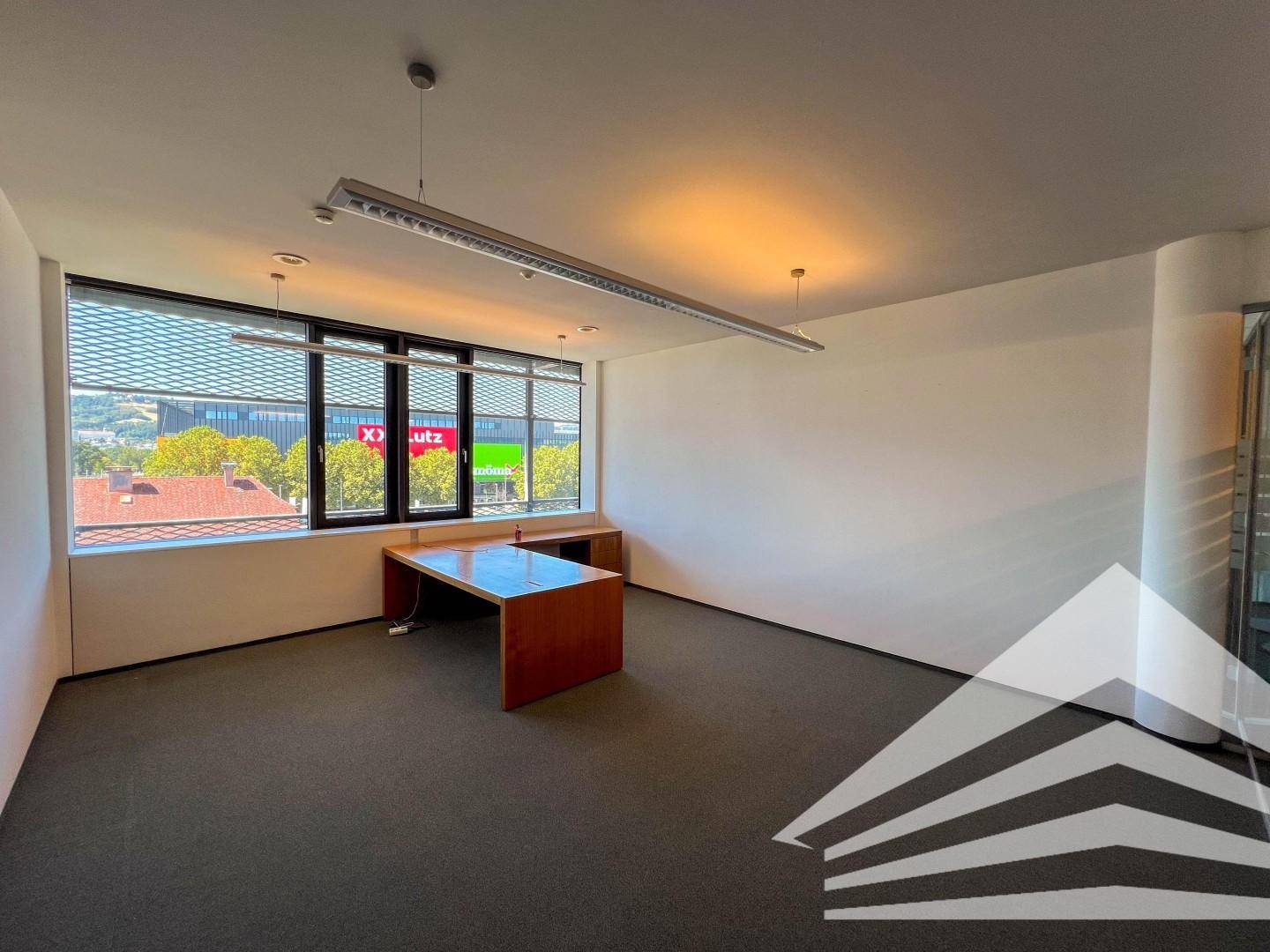 Helles Büro mit Teppichboden, einem Holzschreibtisch und großen Fenstern mit Blick auf die Umgebung.