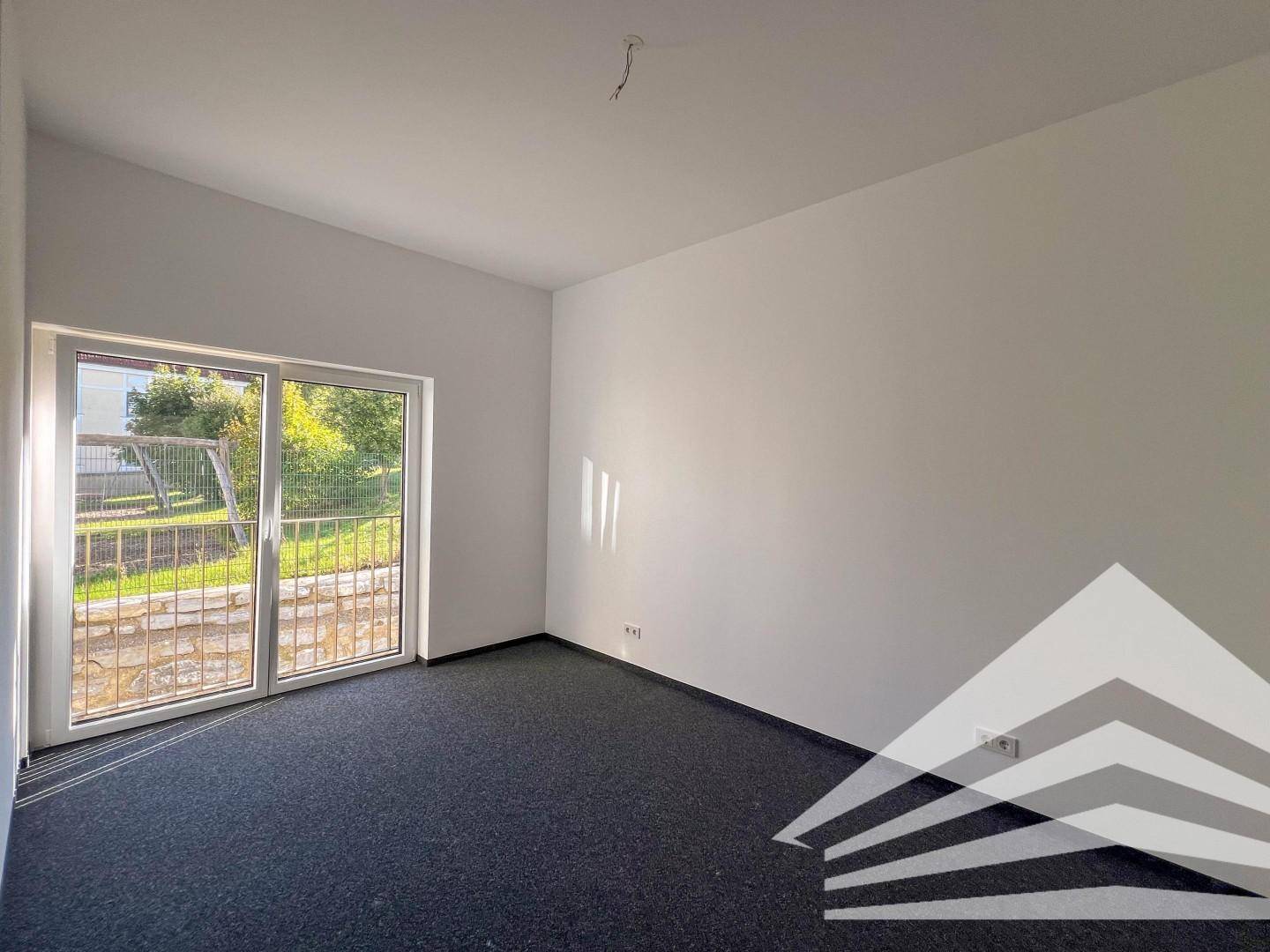 Helles Zimmer mit Teppichboden und großer Schiebetür, die zu einem Außenbereich führt.