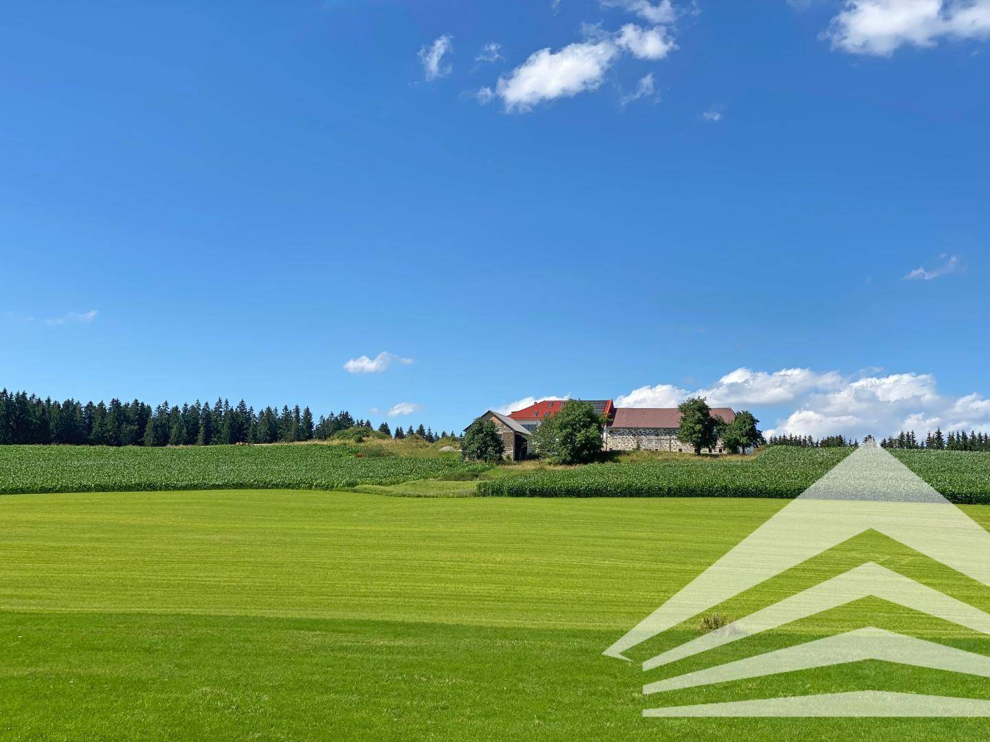 Panoramablick auf einen Gebäudekomplex inmitten weitläufiger grüner Felder unter blauem Himmel.