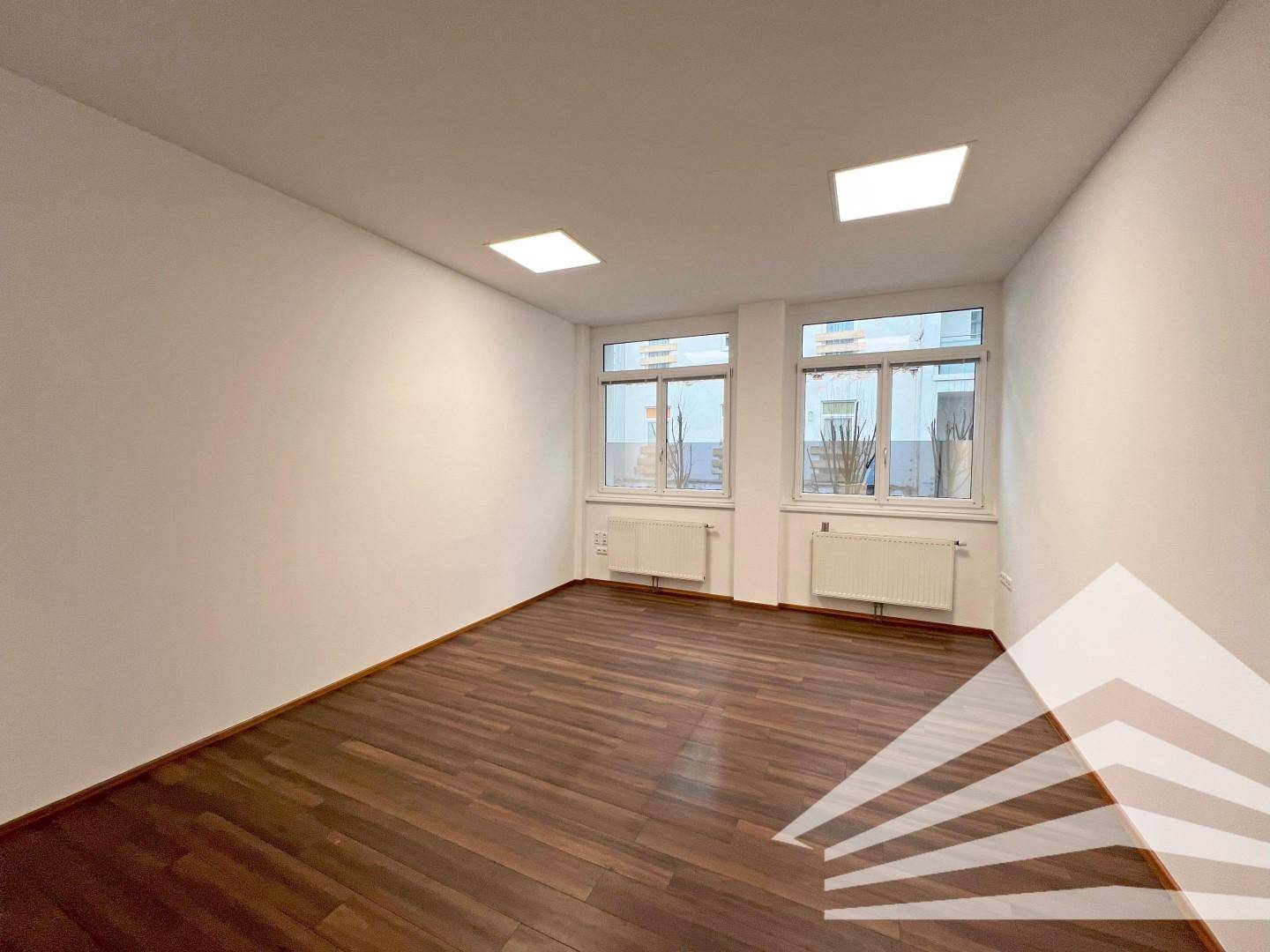 Helles Büro mit zwei Fenstern, Heizkörpern und dunklem Holzboden, bereit zur Nutzung.