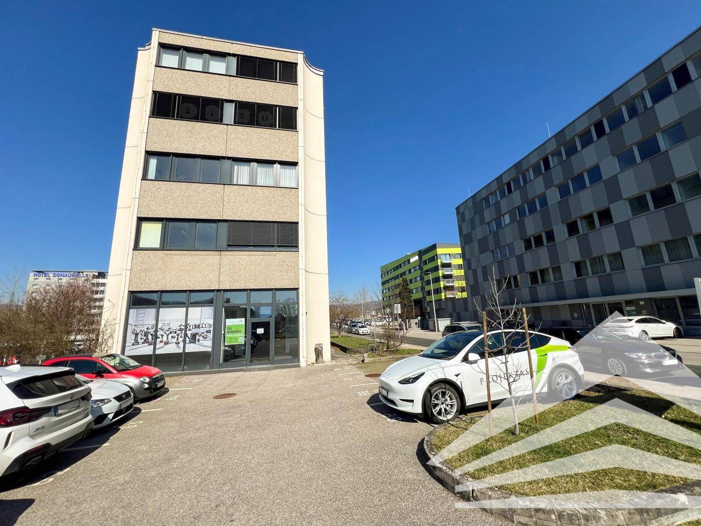 Geschäftsgebäude mit Parkplätzen davor und einem weiteren Gebäude im Hintergrund unter strahlend blauem Himmel.
