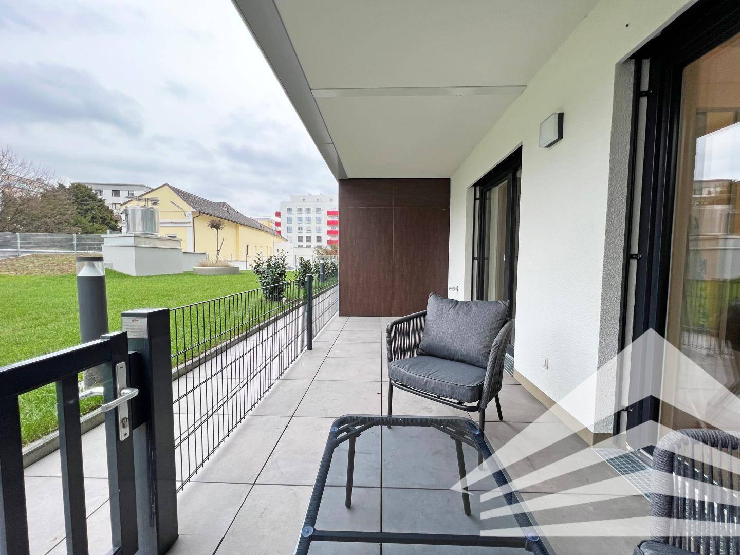 Lange Terrasse mit modernen Fliesen und bequemen Outdoor-Sesseln, umgeben von Grünflächen.