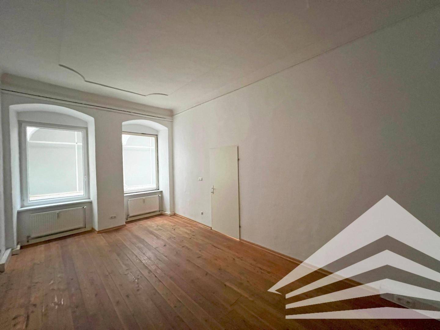 Heller Raum mit zwei hohen Fenstern, Holzdielenboden und einer schlichten, aber eleganten Stuckverzierung an der Decke.