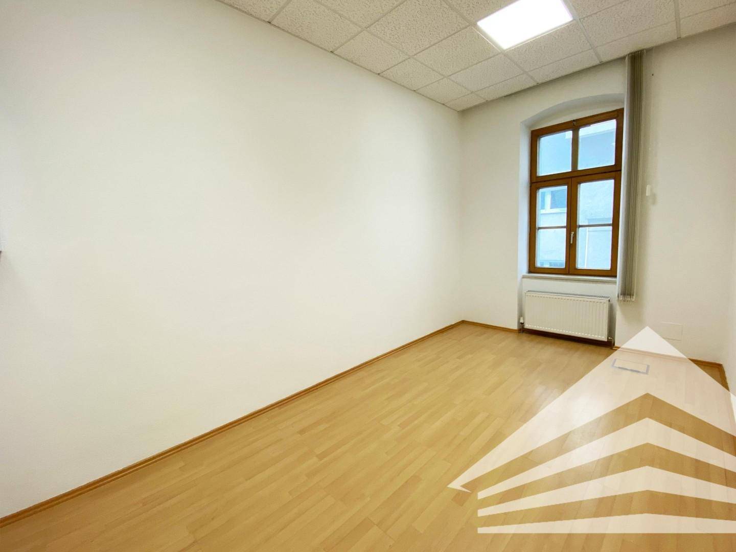 Kleinerer Büroraum mit einem Fenster, hellem Laminatboden und weiß gestrichenen Wänden.