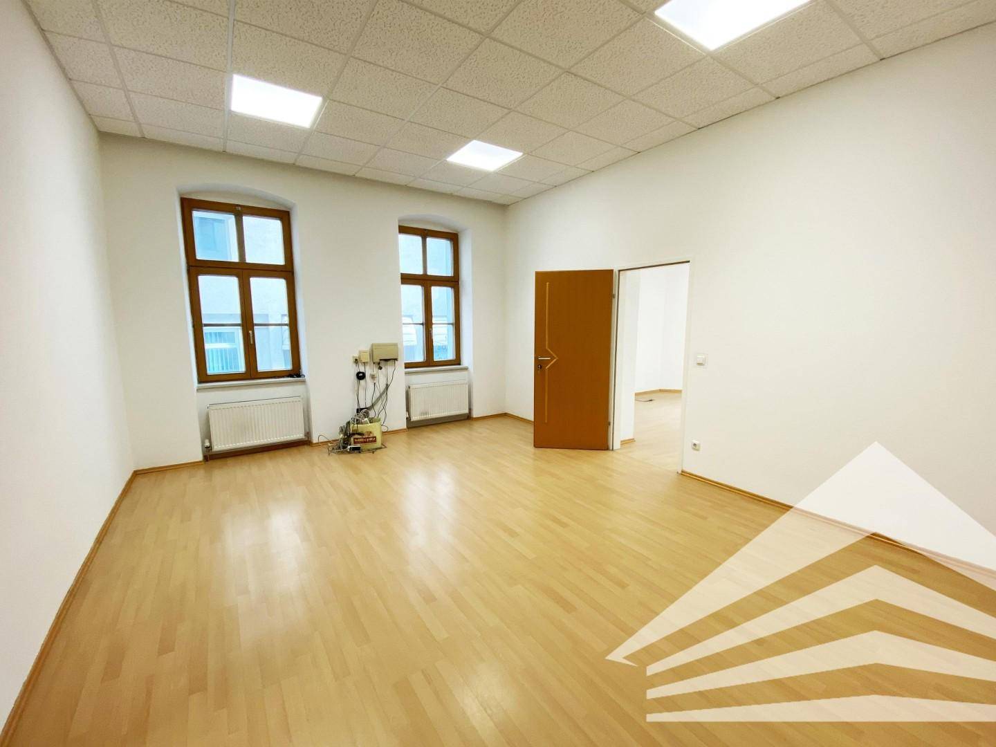 Geräumiger Büroraum mit Holztür, Laminatboden und Blick in einen weiteren Raum.