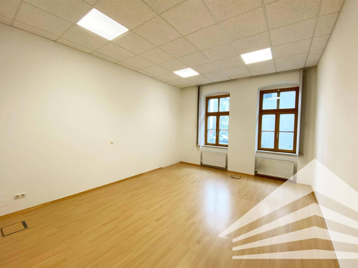 Leerer Büroraum mit zwei Fenstern, hellem Boden und Deckenplatten mit integrierter Beleuchtung.