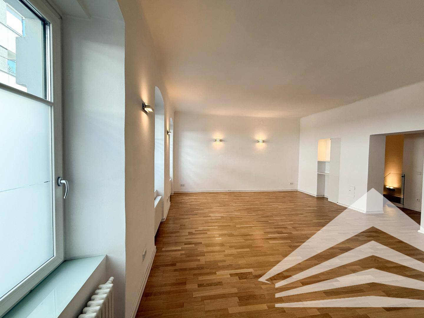 Heller Raum mit Holzboden und einem großen Fenster, bietet Zugang zu einem weiteren Bereich.