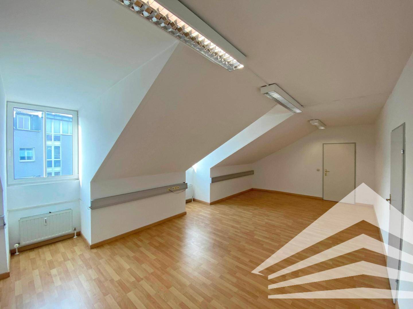Leerer Raum mit Fenster, Heizkörper und Tür, gekennzeichnet durch schräge Dachschrägen und hellen Laminatboden.