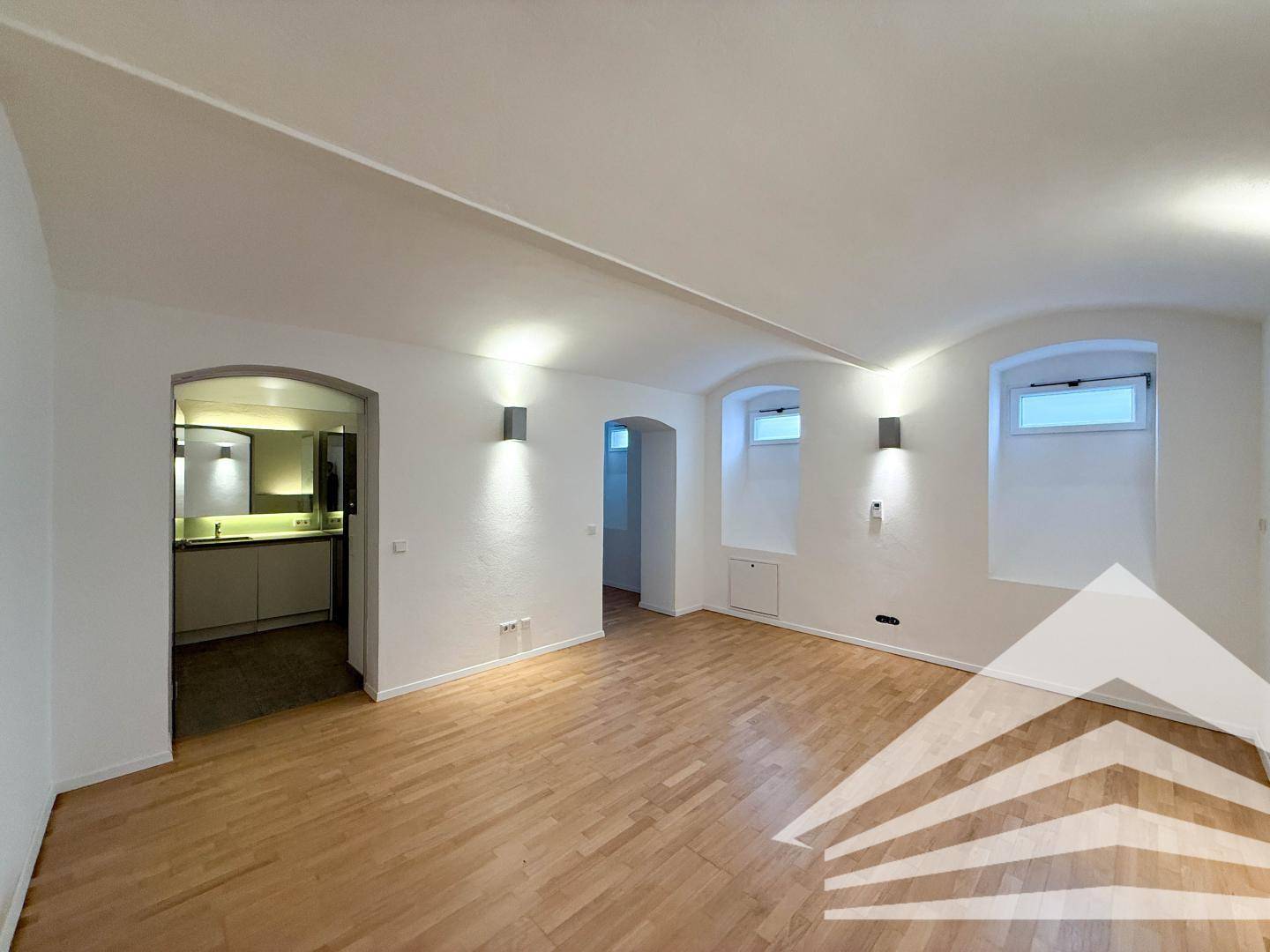 Geräumiger Raum mit Holzboden, weißen Wänden und offenem Zugang zu einer modernen Küche.