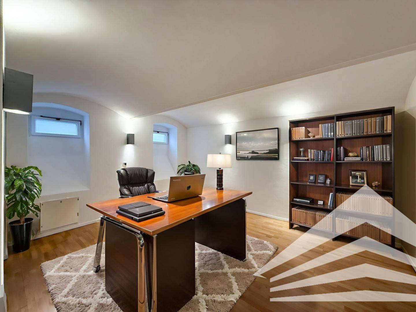 Gemütliches Büro mit Holzboden, Schreibtisch, Bücherregal und zwei kleinen, gewölbten Fenstern.