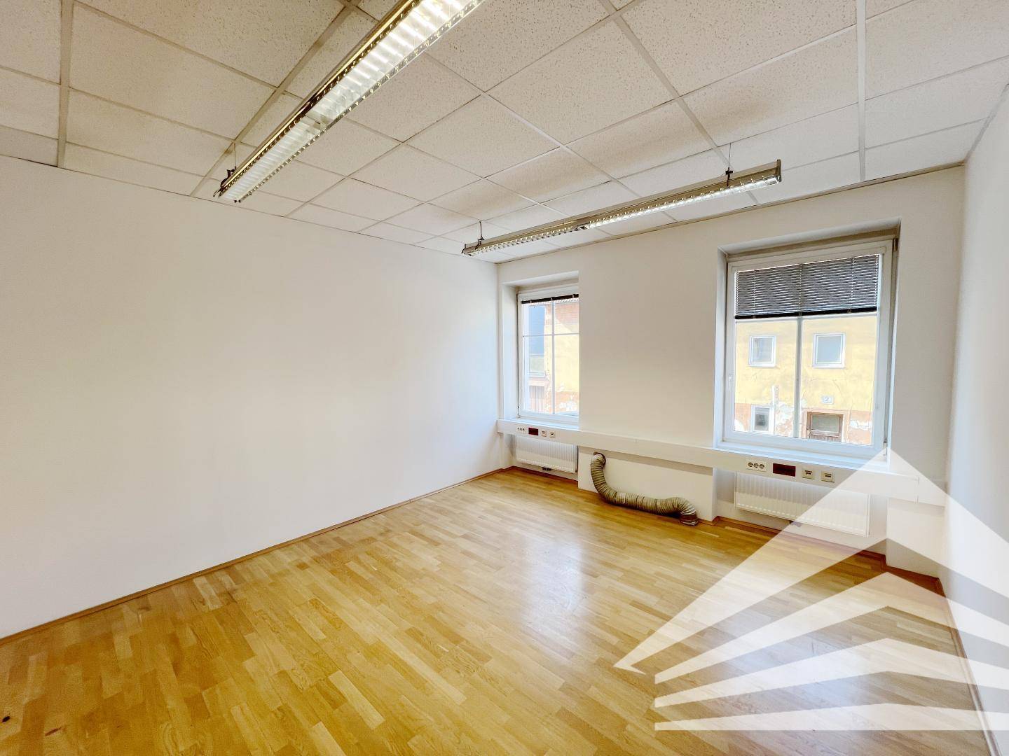 Leeres Büro mit zwei großen Fenstern, die viel Tageslicht hereinlassen, und einem Holzfußboden.