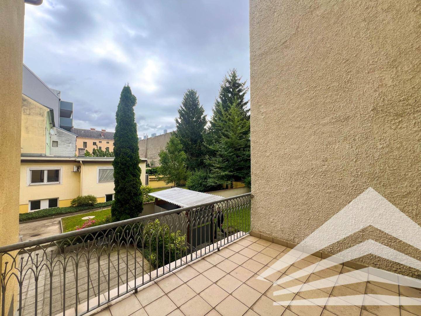Balkon mit schmiedeeisernem Geländer und Blick auf einen grünen, baumbestandenen Innenhof.