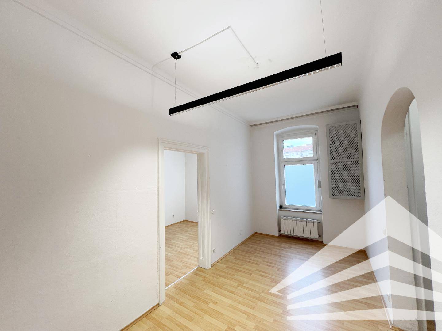 Leerer Raum mit hellem Parkettboden, weißen Wänden und einem Fenster, das viel Tageslicht hereinlässt.