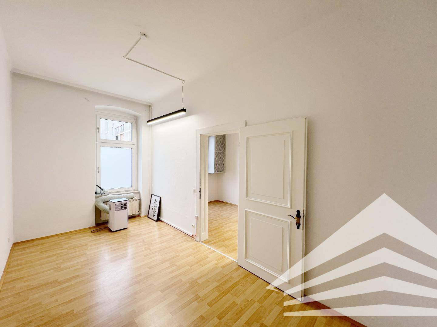 Heller Büroraum mit Holzboden, großem Fenster und einer Tür, die zu einem weiteren Zimmer führt.