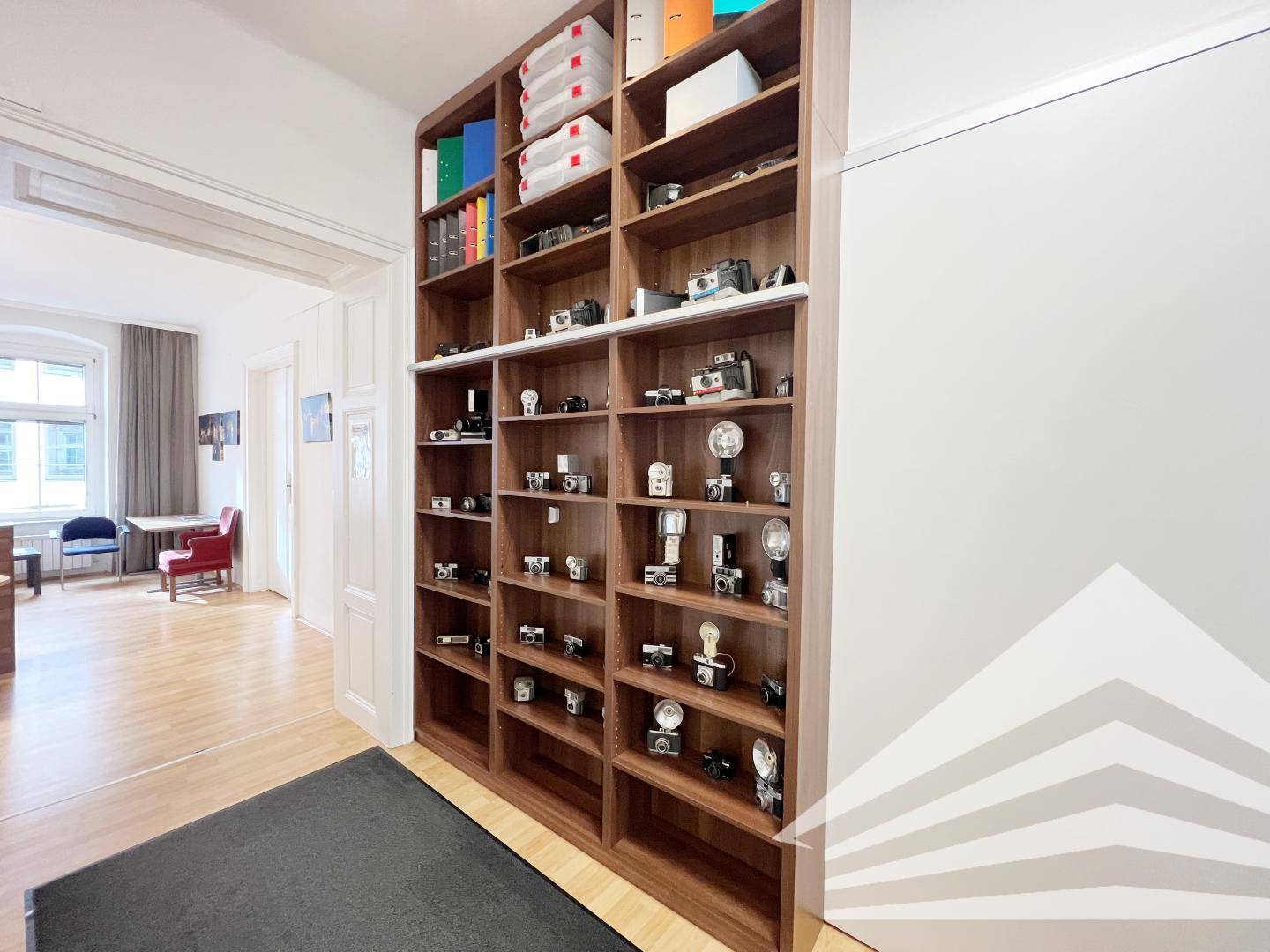 Hohes Holzregal mit einer Sammlung von Kameras und Büchern, im Hintergrund ist der Bürobereich sichtbar.