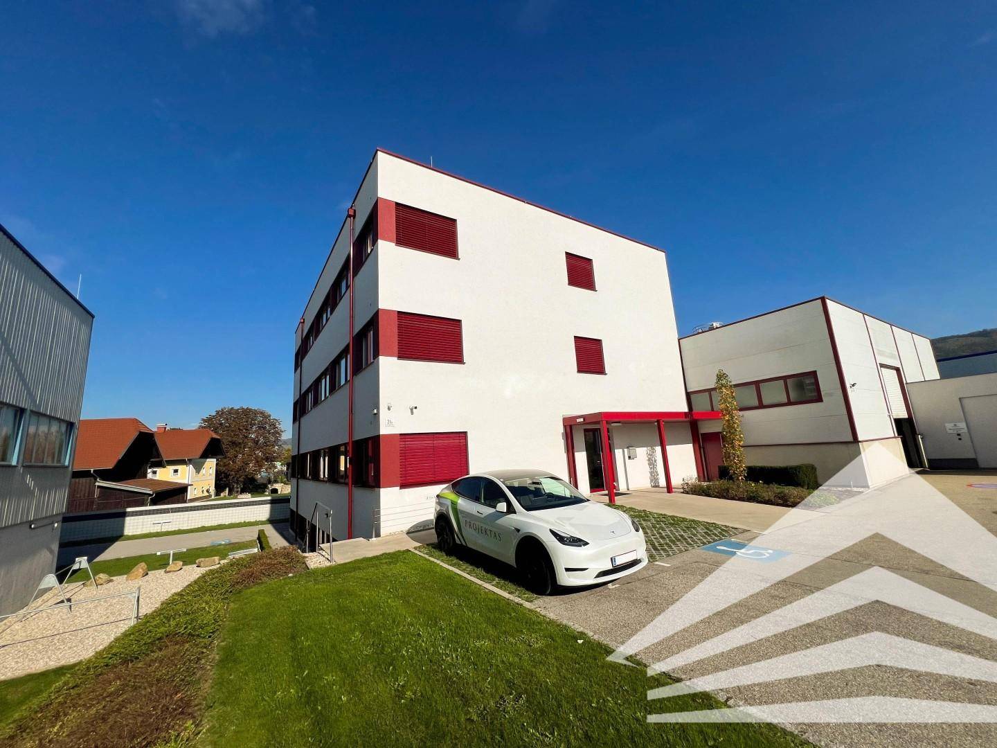 Modernes Bürogebäude mit weißer Fassade, roten Akzenten und einem geparkten Elektrofahrzeug auf dem Vorplatz.