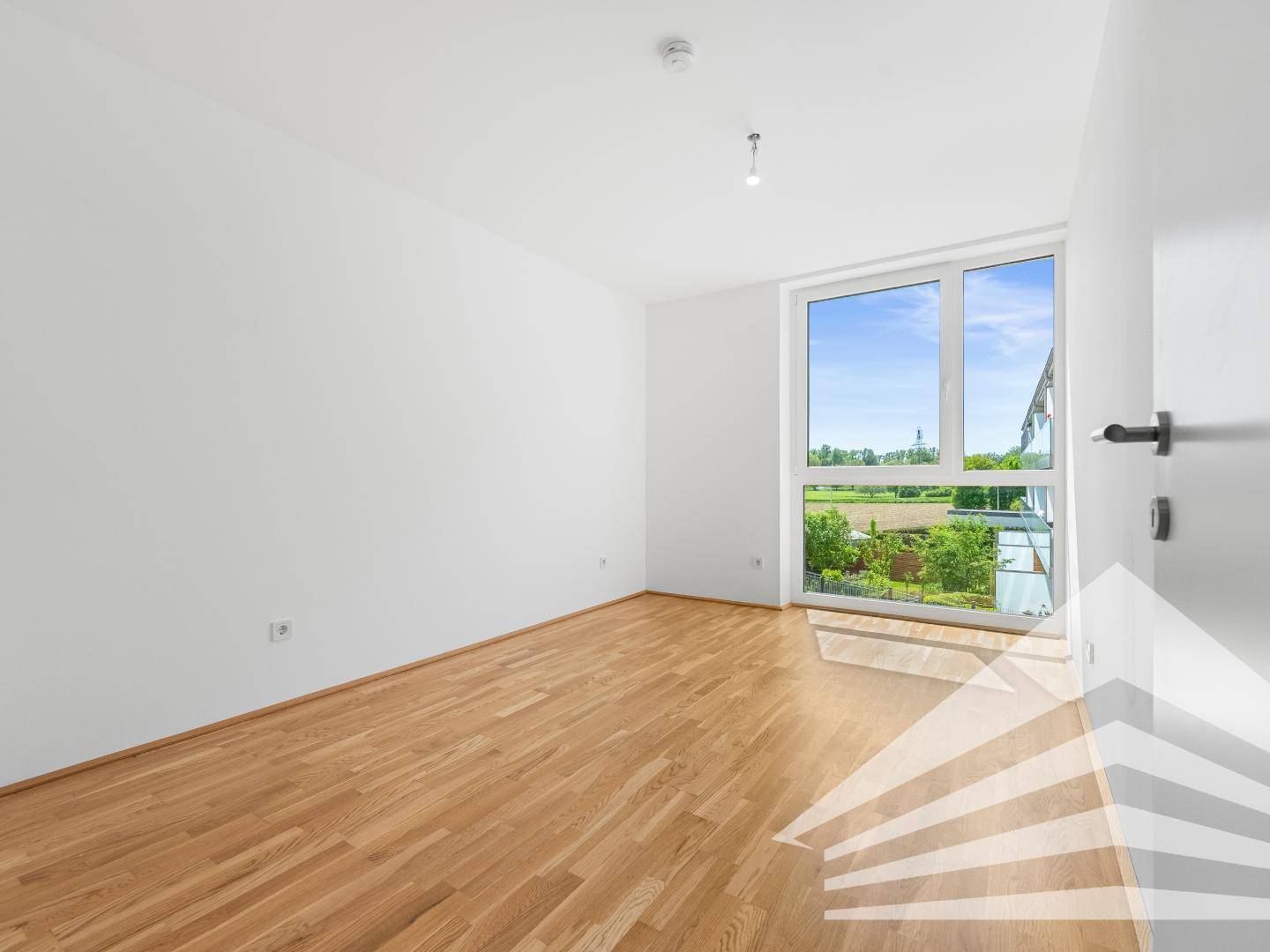Leerer Raum mit Parkettboden und großem Fenster, das viel Tageslicht hereinlässt.