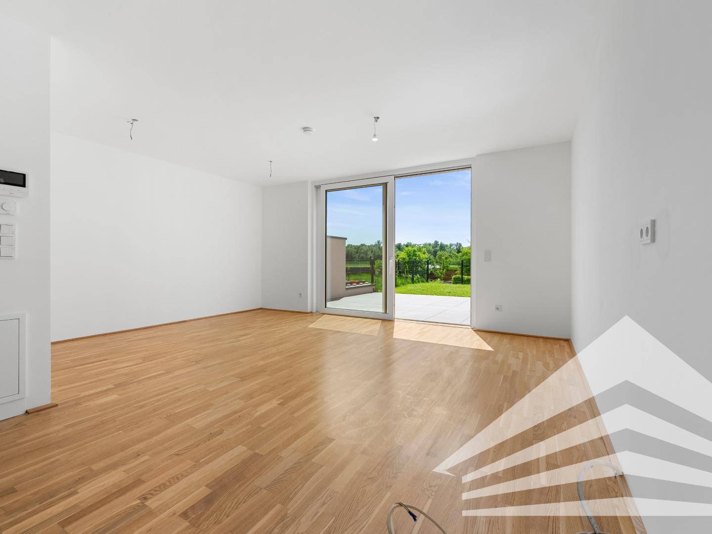 Helles, leeres Wohnzimmer mit Parkettboden und Zugang zur Terrasse mit Grünblick.