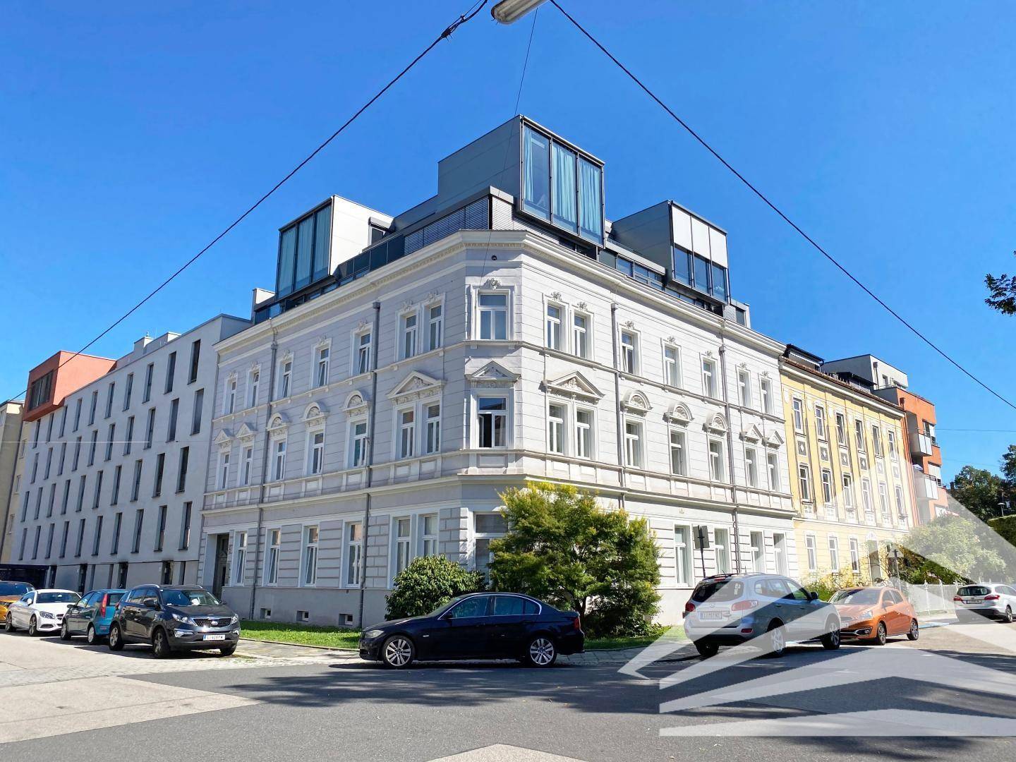 Historisches Gebäude mit modernen Dachaufbauten an einer städtischen Straße mit parkenden Autos.