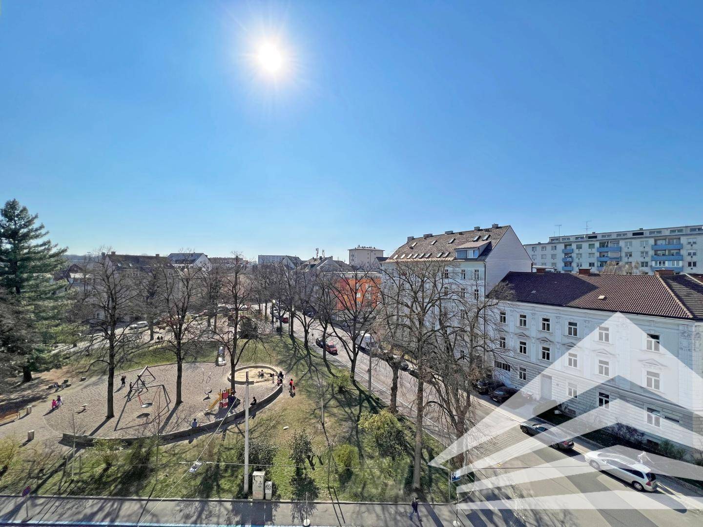 Blick auf einen sonnigen Stadtpark mit Spielplatz und umliegenden Gebäuden, ideal für Familien.