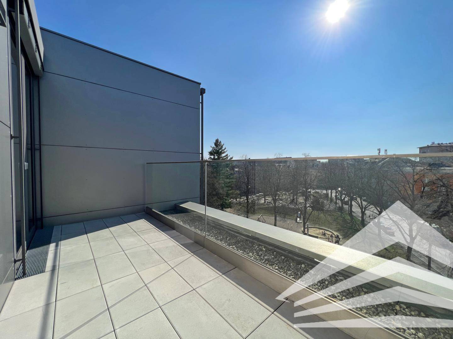 Sonnige Terrasse mit Fliesenboden und Glasgeländer, bietet einen Blick auf den Park und die Stadt.