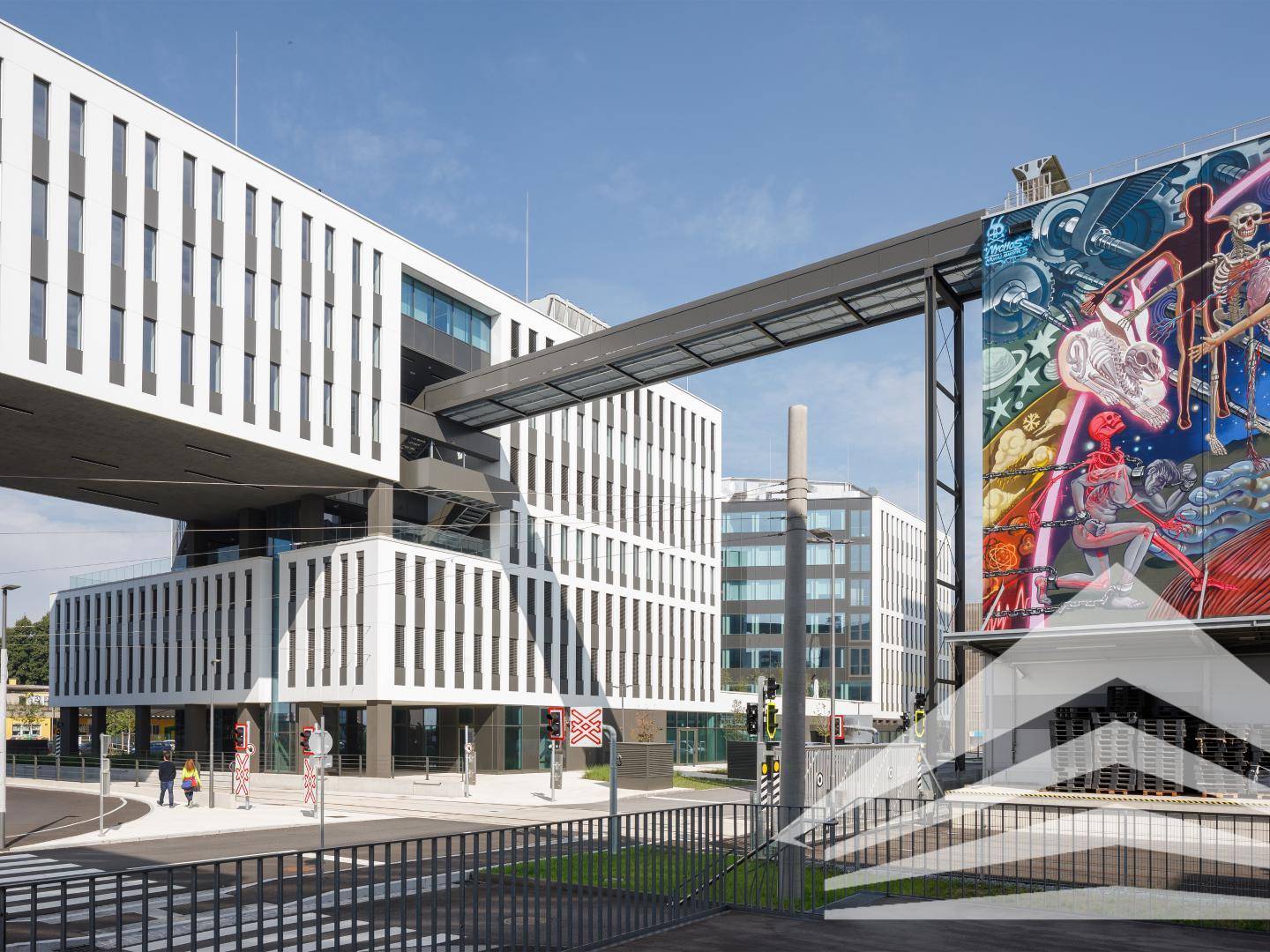 Moderner Gebäudekomplex mit einer Verbindungsbrücke und einem großen, farbenfrohen Wandgemälde, das Kunst und Architektur verbindet.
