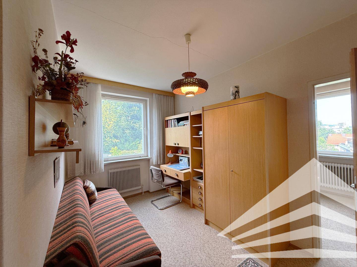 Helles Zimmer mit Teppichboden, Schreibtisch und Fenster mit Blick ins Grüne.