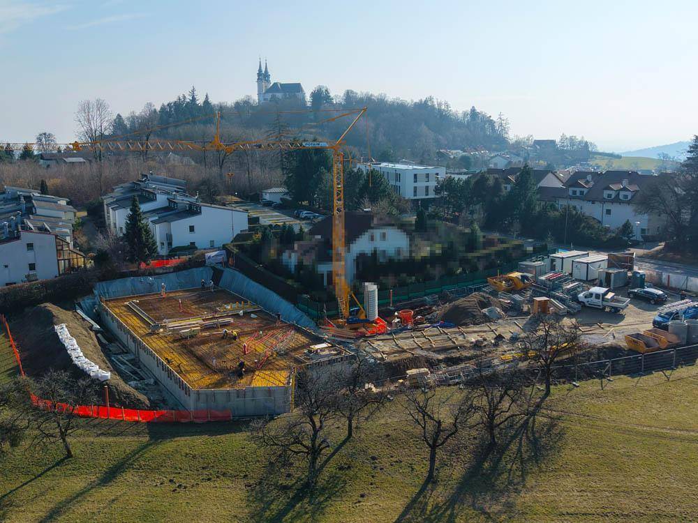 Nahaufnahme der Baustelle mit dem Fundament des Neubaus und einem Baukran vor einer hügeligen Landschaft.