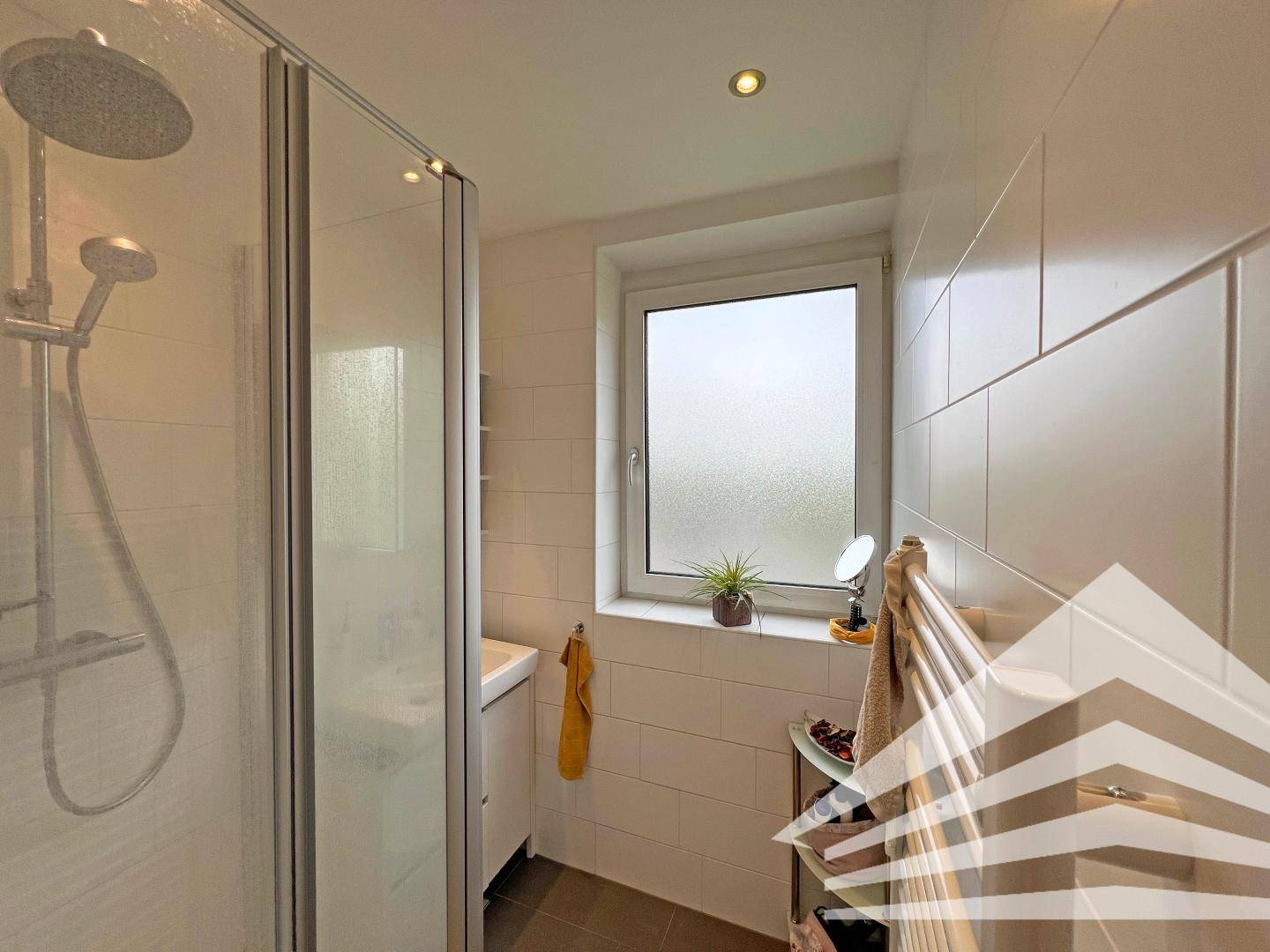 Modernes Badezimmer mit Duschkabine, Waschbecken und Fenster, hell gefliest.