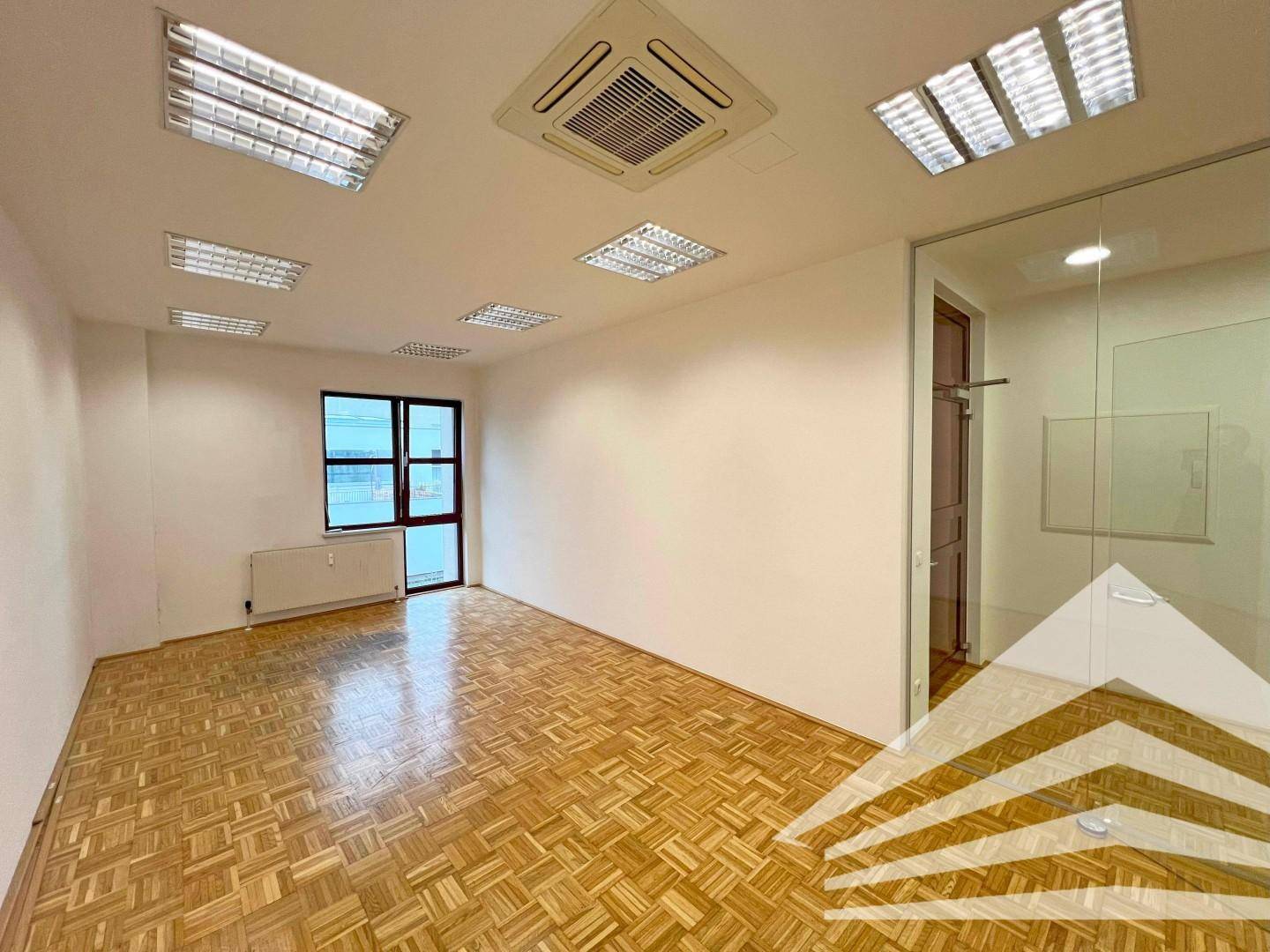 Leerer Büroraum mit Parkettboden, einem Fenster und Zugang zu einem weiteren Bereich.