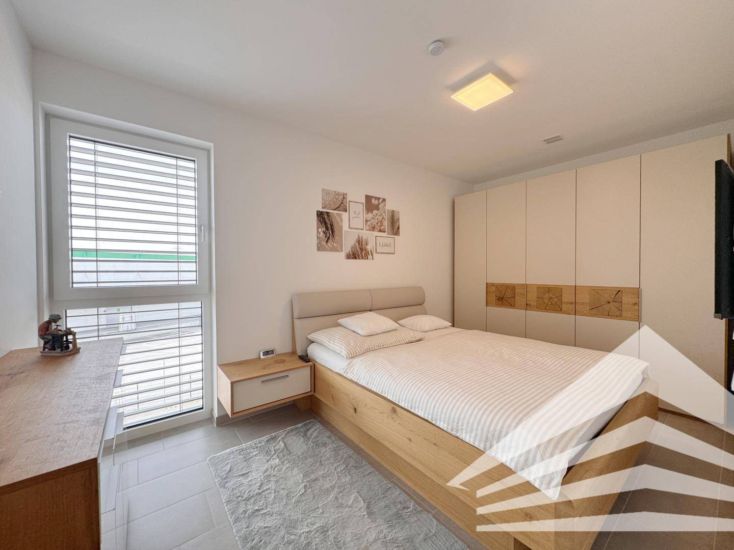 Helles Schlafzimmer mit Doppelbett, Einbauschrank und großem Fenster mit Jalousien.