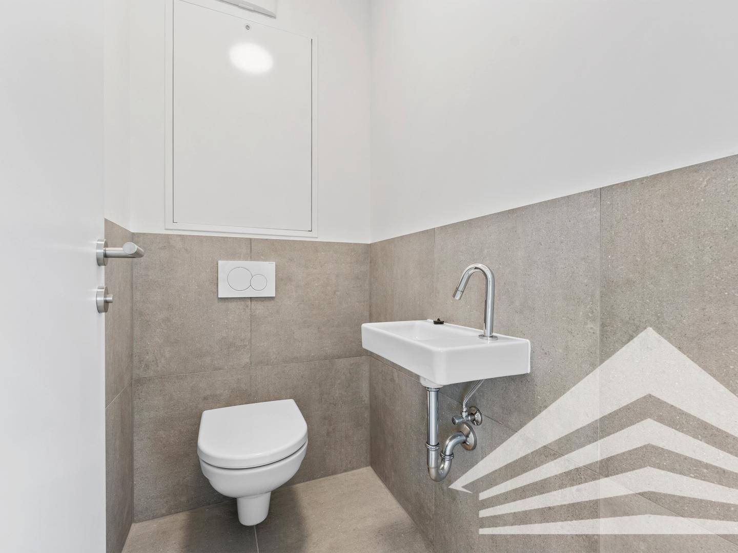 Separates WC mit wandhängender Toilette, kleinem Waschbecken und grauen Fliesen.