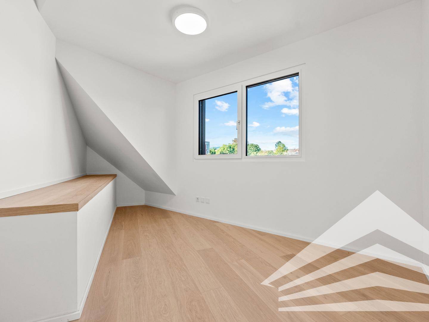 Heller Dachraum mit schräger Decke, Parkettboden und großem Fenster mit Ausblick.