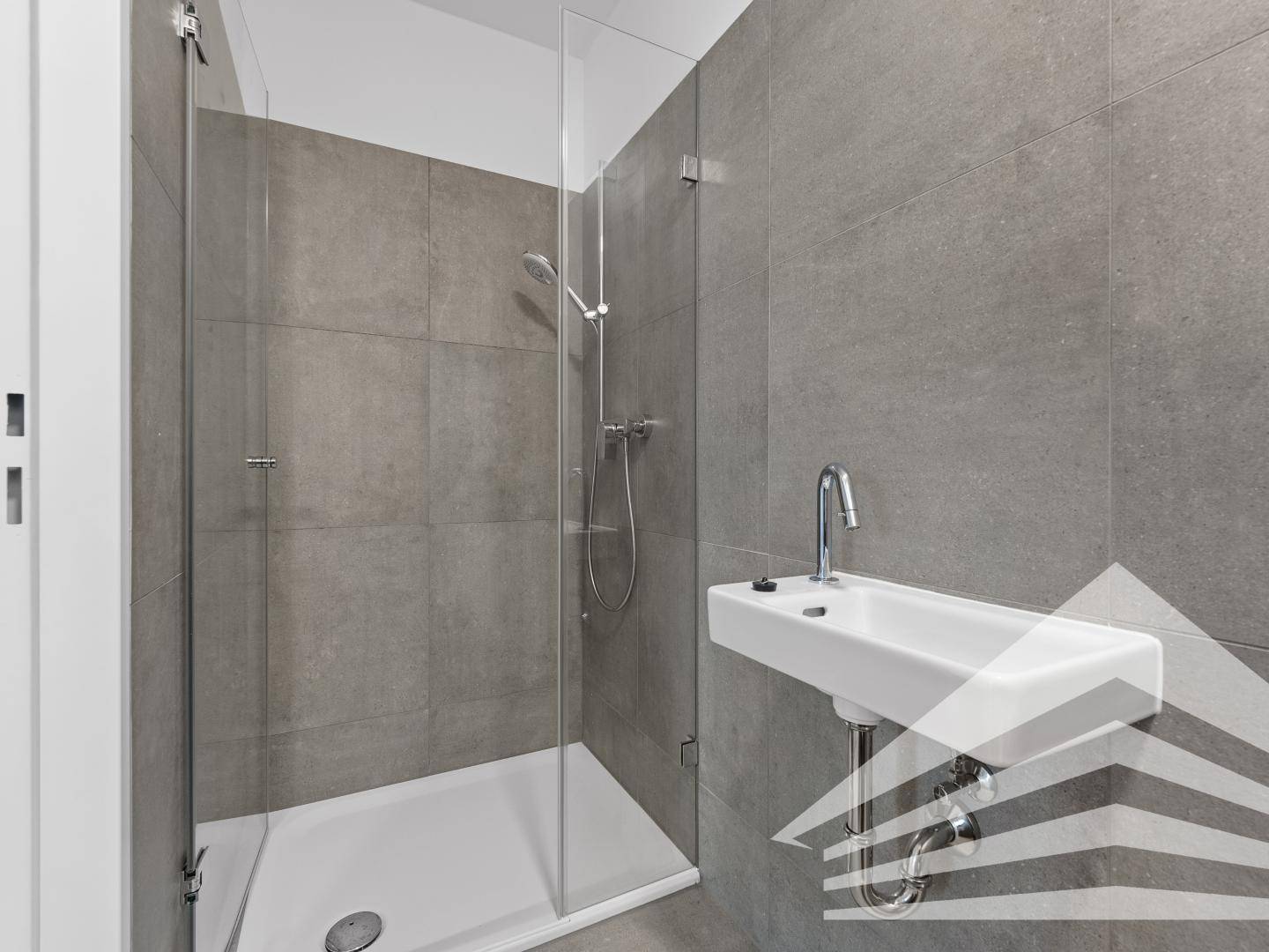 Modernes Badezimmer mit großen grauen Fliesen, ebenerdiger Dusche und minimalistischem Waschbecken.