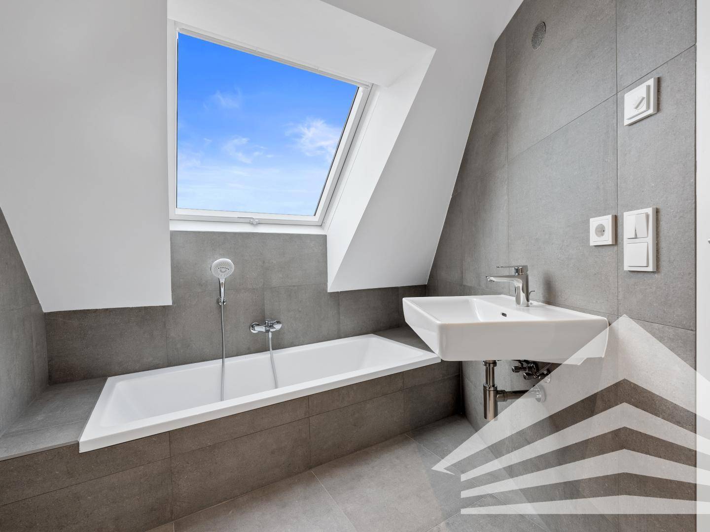 Modernes Badezimmer mit Badewanne, Waschbecken und Dachfenster, in hellen Grautönen gehalten.