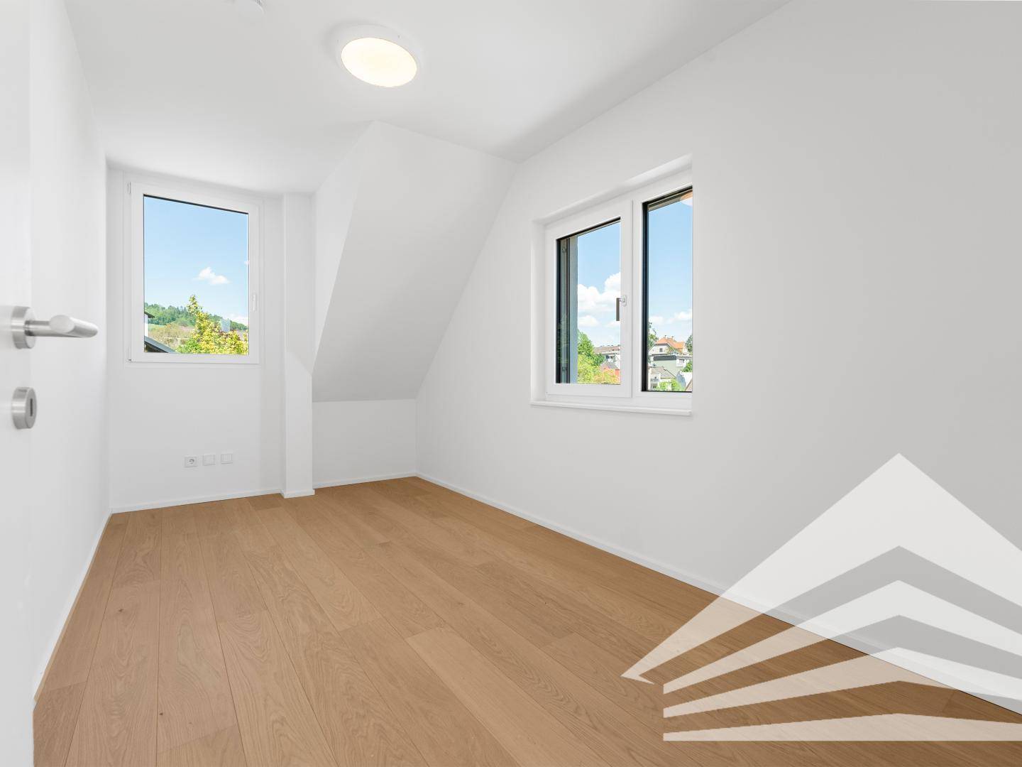 Helles Zimmer mit zwei Fenstern, Holzboden und weiß gestrichenen Wänden.