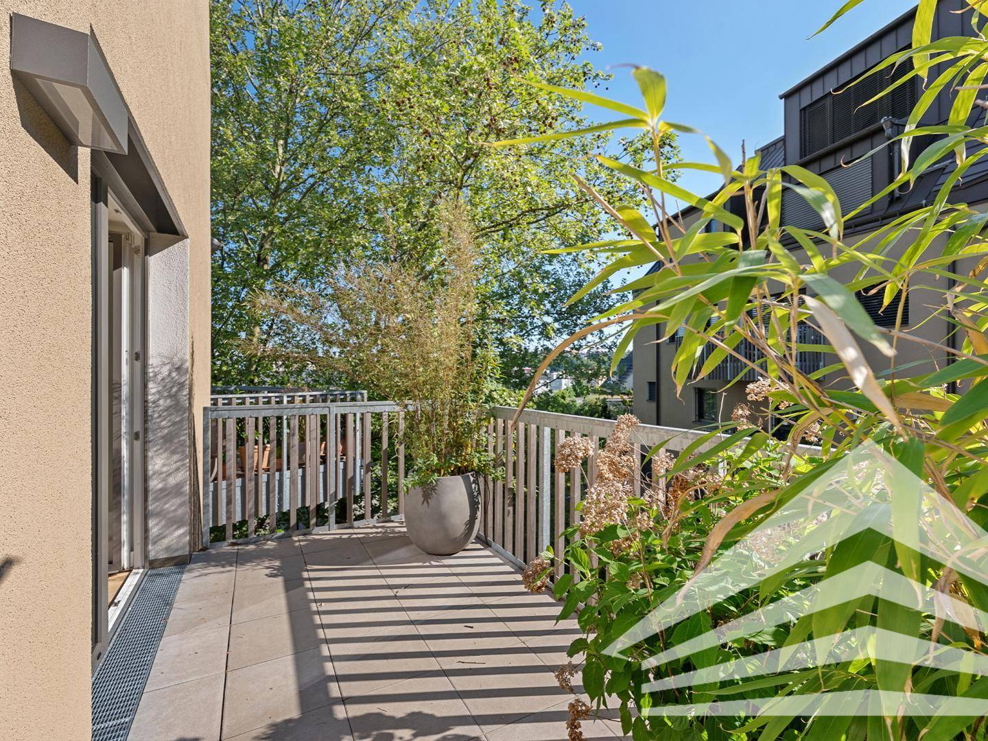 Sonniger Balkon mit Fliesenboden, Pflanzen und Blick auf grüne Bäume und umliegende Gebäude.