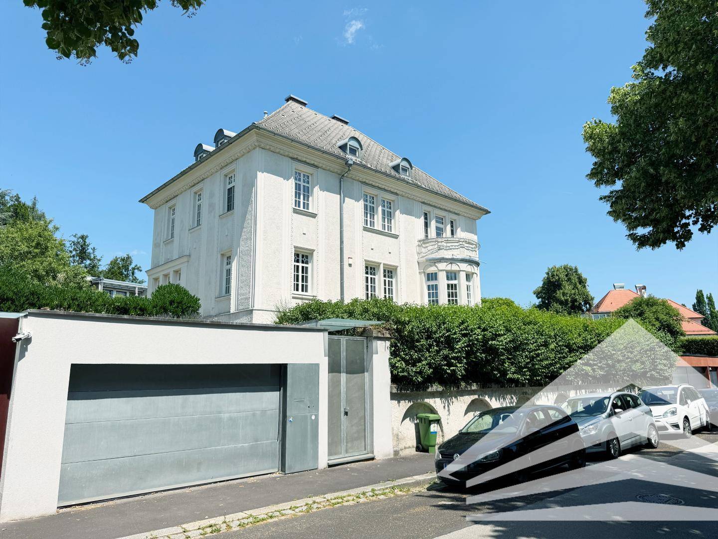 Villa von der Straße aus gesehen mit Garage, Hecke und parkenden Autos.
