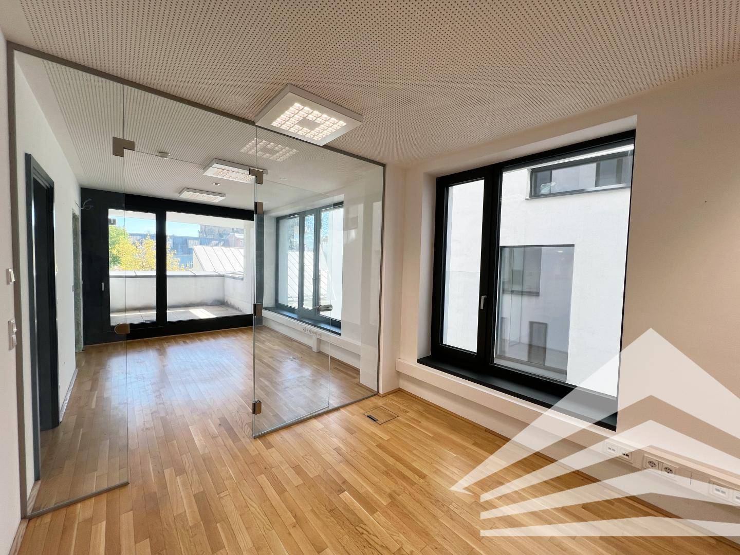 Moderner Büroraum mit Holzboden, großen Fenstern und einer eleganten Glaswand als Raumteiler.