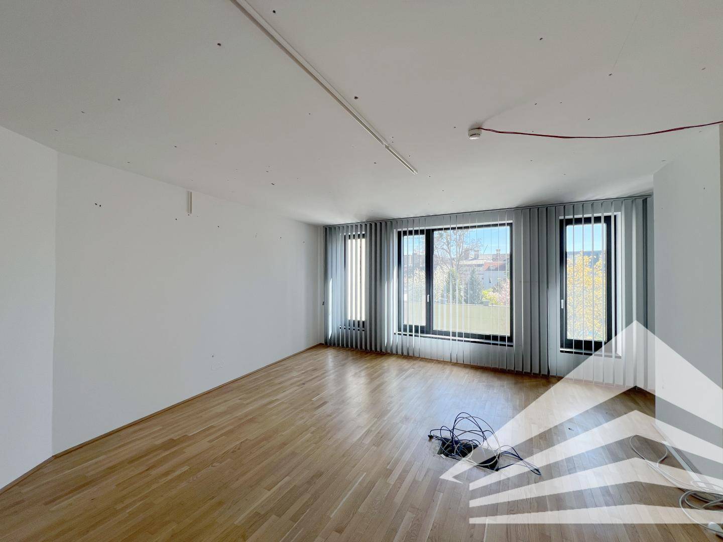 Leerer Büroraum mit großen Fenstern, Jalousien und Holzboden, bereit zur Einrichtung.
