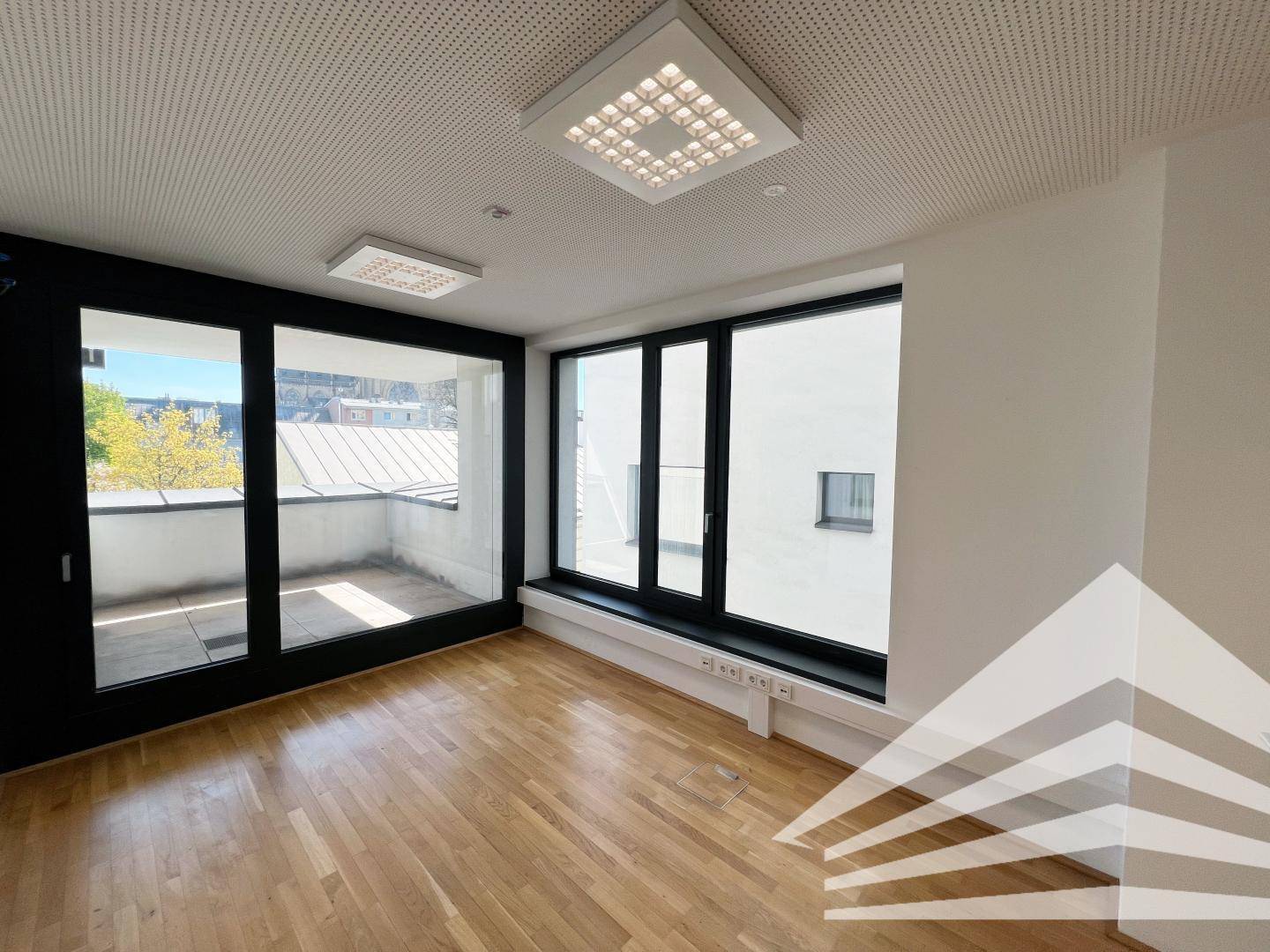 Heller Raum mit Holzboden, großen Fenstern und Zugang zu einem Balkon, ideal für Büronutzung.